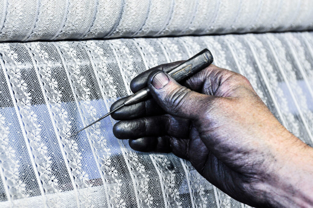  - Atelier Leavers de l'usine Codentel.  Main de tulliste noircie par le graphite qui enduit les chariots des métiers Leavers. Avec son crochet, fabriqué à partir d'une baleine de parapluie, il démêle les fils flottants du motif sur le tissage en cours. Codentel a été repris par le groupe Holesco en 2016.