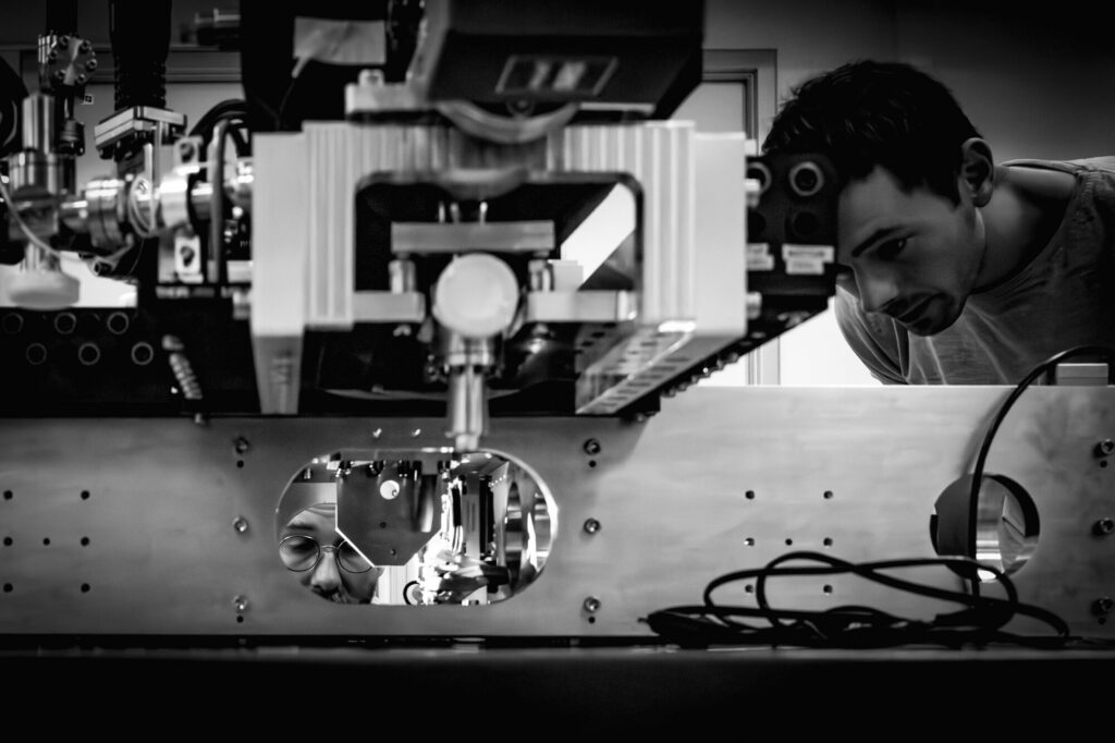 Be Monochrom - Photo 2 : À hauteur de machine Des ingénieurs observent et ajustent un dispositif quantique développé par PASQAL. La photographie montre la précision des opérations nécessaires à la mise au point de cette technologie de pointe, entre maîtrise instrumentale et attention humaine.