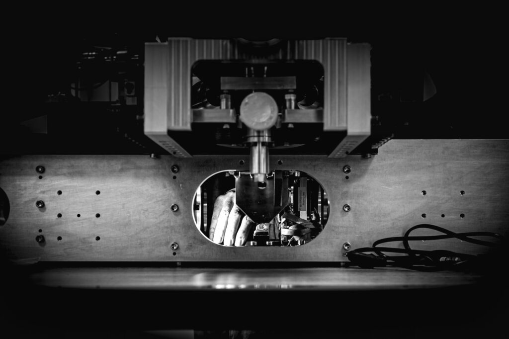 Be Monochrom - Photo 3 :L’intelligence de la main Une main intervient à l’intérieur d’un dispositif quantique lors d’une opération de réglage. L’image souligne la dimension concrète et sensible d’une technologie souvent perçue comme abstraite, où le geste humain reste essentiel.
