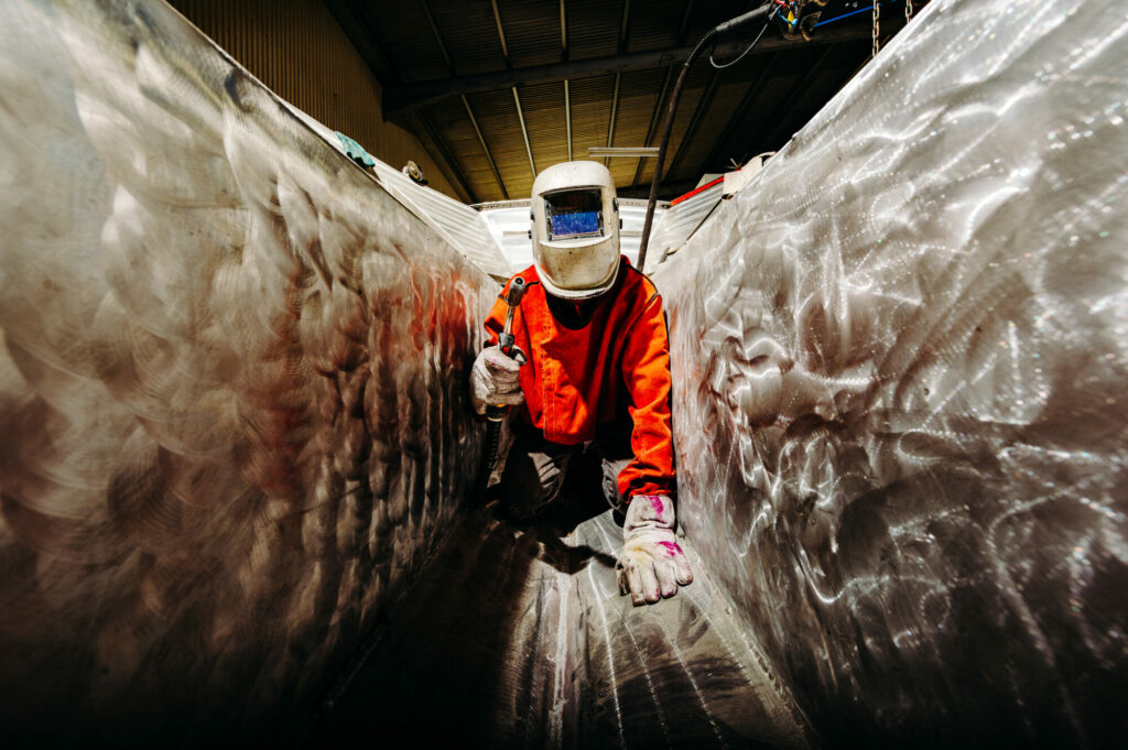 Lumen33 - Précision et savoir-faire naval : Un soudeur expert de Nauticalu Nouvelle-Calédonie, parfaitement équipé en EPI (masque, veste, gants), est concentré sur l'assemblage d'une structure en aluminium à l'intérieur d'une coque de bateau en construction.